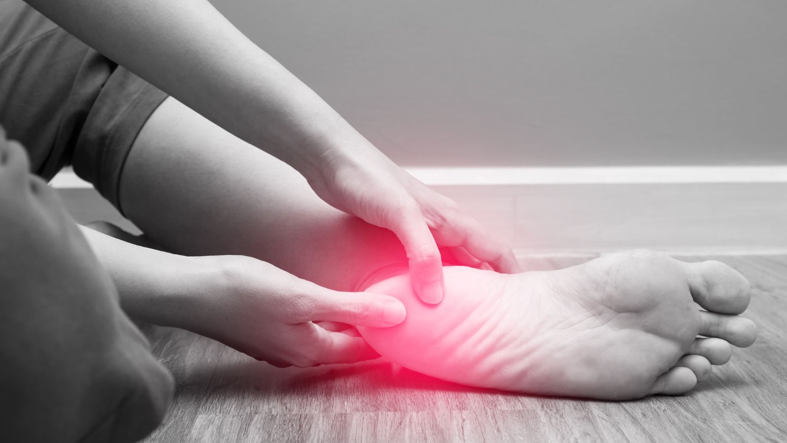 A woman sits on the ground with her hand pressing down on her heel, which is painful due to plantar fasciitis.