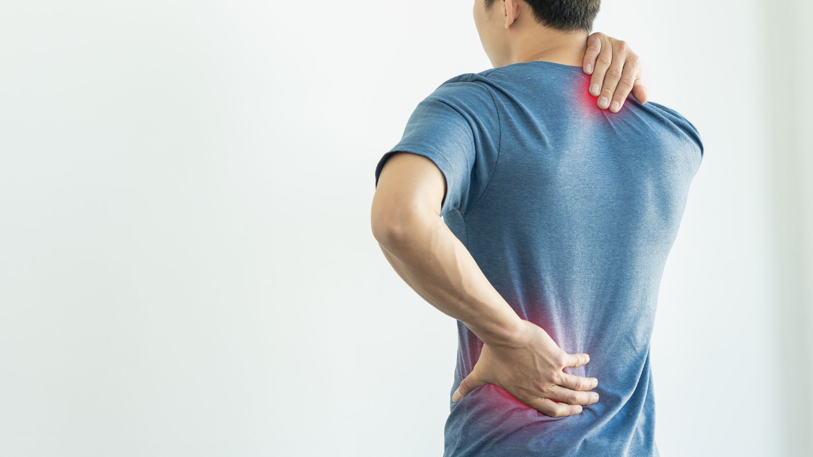 A young man feeling discomfort due to lower back pain is using his hands to massage his shoulders and lower back.