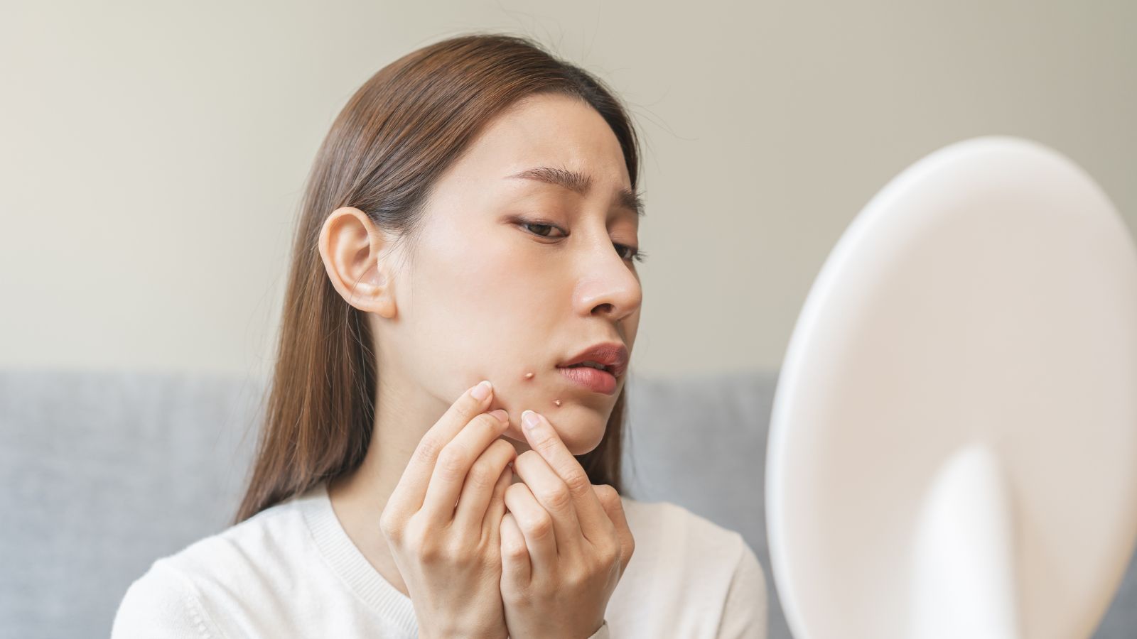 An Asian woman expresses concern as she touches acne around her chin and mouth, contemplating squeezing the acne spots on her face.